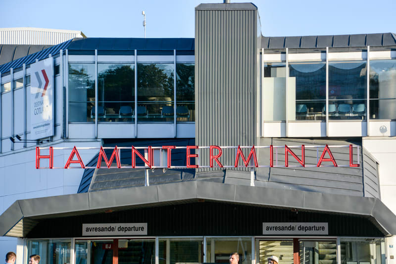 Bilden visar fasaden på en byggnad med texten "HAMNTERMINAL" i röda neonbokstäver centrerad ovanför ingångspartiet. Byggnaden har stora fönsterpartier på övervåningen som reflekterar omgivande träd och himmel. Innanför fönstren syns möbler och stolar