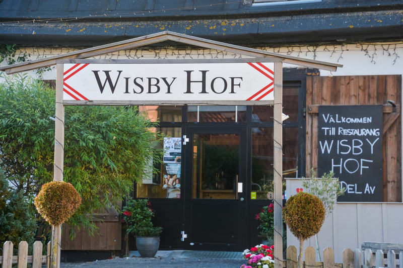 En bild som visar en skylt för "WISBY HOF" med röd-vita chevronmönster på sidorna, monterad på en träram som bildar en portal. Under skylten finns en svart dörr flankerad av fönster. Till vänster om dörren står en grön buske och vid dörrens nederkant