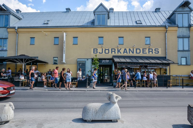 En bild tagen utomhus under dagtid, föreställandes en gatuscen med en gul byggnad i bakgrunden. Byggnaden har texten "BJÖRKANDERS" på fasaden. Framför byggnaden finns flera uteserveringar med människor sittande vid bord och i en bar. Några personer g