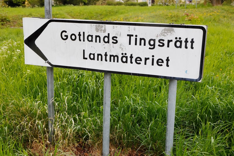 En vit vägskylt med svarta bokstäver och en svart vänsterpekande pil är placerad i bildens nedre vänstra hörn. Skylten är fäst vid två grå metallstolpar. Texten på skylten lyder "Gotlands Tingsrätt Lantmäteriet". Bakgrunden består av grön gräsmark me