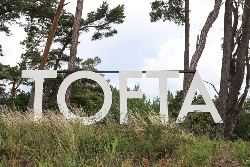 En stor, vit skylt med bokstäverna "TOFTA" är placerad utomhus. Under skylten växer högt, torrt gräs och annan grön vegetation. Bakom skylten och gräset syns en tät skog med barrträd, främst tallar, med grova stammar och grönskande barr. Himlen ovanf