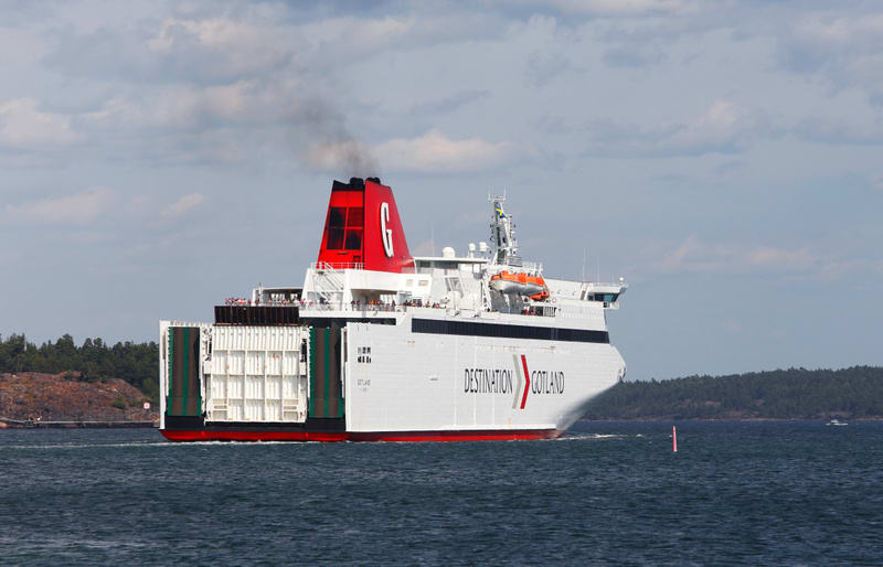 En vit färja med röd botten och en röd skorsten med ett stort vitt 'G' på, rör sig från vänster till höger i bild på ett blågrönt vatten. Färjan har texten 'DESTINATION GOTLAND' längs sidan. Från skorstenen stiger svart rök. Aktern på färjan är öppen