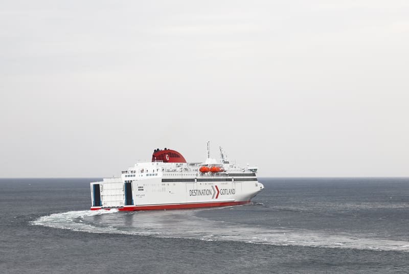 Bilden visar ett stort passagerarfartyg, en färja, i rörelse på öppet vatten. Fartyget är vitt med en röd linje längs skrovet och en röd skorsten med ett vitt 'G'. På fartygets sida står texten "DESTINATION GOTLAND" med pilar. Aktern av fartyget, med