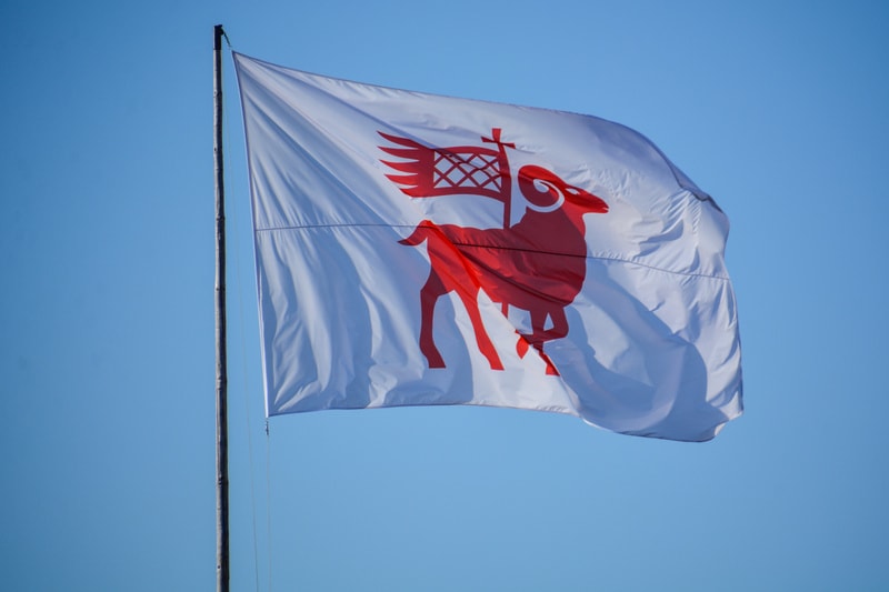 En vit flagga med ett rött motiv av en bagge syns vajandes. Baggen står upprätt och håller en röd flagga på en kort stång. Baggen har horn och detaljer som antyder päls. Bakgrunden består av en klarblå himmel. Flaggan är fäst vid en lodrät flaggstång