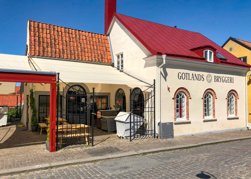 En utomhusbild tagen under dagtid som visar en byggnad med texten "GOTLANDS BRYGGERI" på fasaden. Byggnaden har en ljus fasad och ett rött plåttak med en röd skorsten. På den vänstra delen av byggnaden finns en terrass med ett ljust tak av tyg, en rö