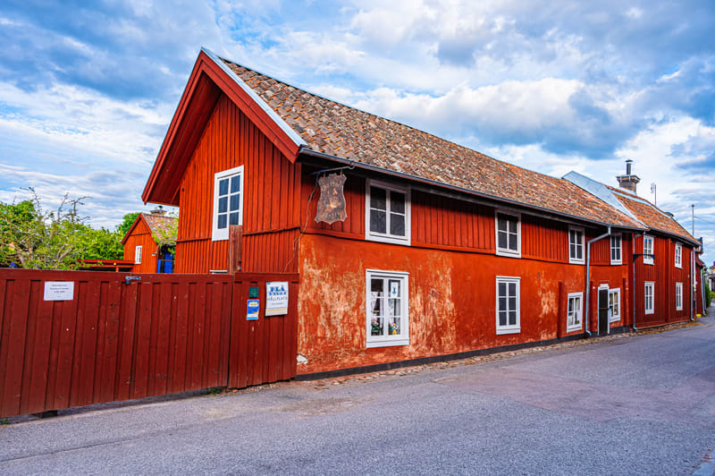 Bilden visar en gata med flera röda trähus under en molnig himmel. I förgrunden till vänster syns ett högt rött trästaket som sträcker sig längs gatan. Bakom staketet skymtar grönskande träd och buskar. Det närmaste huset är målat i en ljusare röd fä