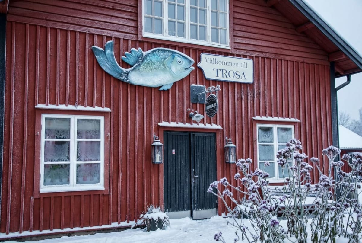 En röd träbyggnad i vinterlandskap. Fasaden är målad i falurött med vita fönsterkarmar och svarta dubbelportar i mitten. Ovanför dörrarna hänger en stor, ljusblå fiskprydnad, och bredvid den sitter en skylt med texten "Välkommen till TROSA". Över dörren hänger även en mindre svart skylt med vit text. Marken framför byggnaden är täckt av snö, och snö ligger även på fönsterbrädorna, takfot och enstaka växter i förgrunden till höger. Fönsterrutorna uppvisar frostmönster. Två lyktor hänger på vardera sida om dubbelportarna.