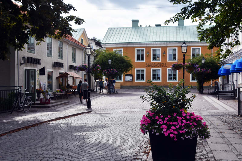 Bilden visar ett torg eller en kullerstensbelagd gata i en stadsmiljö. I förgrunden till höger står en stor svart blomlåda fylld med rosa och vita blommor. Till vänster i bilden syns en vit byggnad med skyltar som innehåller text, bland annat "KIOSK"