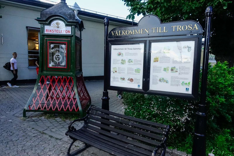 Bilden visar ett utomhusområde med en grönmålad 'RIKSTELEFON'-telefonkiosk med rött gallerverk vid basen. Bredvid telefonkiosken står en svart anslagstavla med texten 'VÄLKOMMEN TILL TROSA' överst. Anslagstavlan innehåller information om Trosa kommun