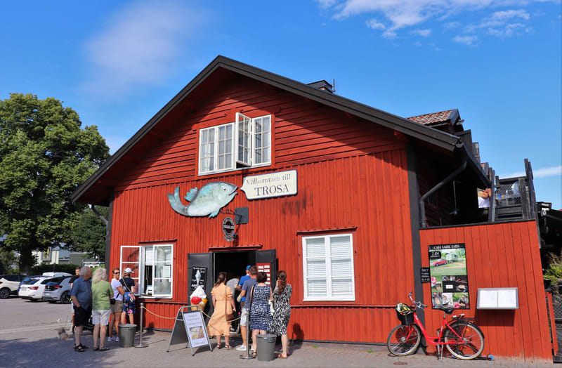 Bilden visar fasaden på ett tvåvåningshus målat i rött med svarta detaljer. På husets framsida finns flera fönster med vita karmar, varav ett på andra våningen står öppet. En stor skylt i form av en blå fisk är monterad på väggen, med en vit textruta