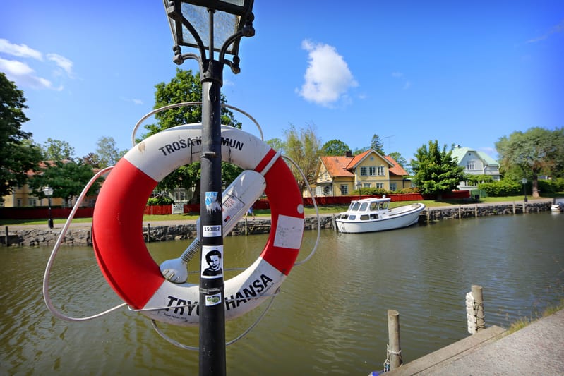 Bilden visar en kanal eller å med en livboj hängandes på en svart lampstolpe i förgrunden. Livbojen är röd och vit och har texten "Trosa Kommun" och "Trygg Hansa" skrivet på sig. På den svarta lampstolpen sitter flera klistermärken, bland annat en me