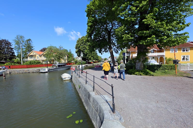 Bilden visar ett landskap med en kanal eller å på vänster sida och en grusgång med träd och hus på höger sida. I vattnet ligger flera båtar, inklusive en större gråfärgad båt och en mindre vit båt. Längs vattnet löper en stenkant med ett svart räcke.