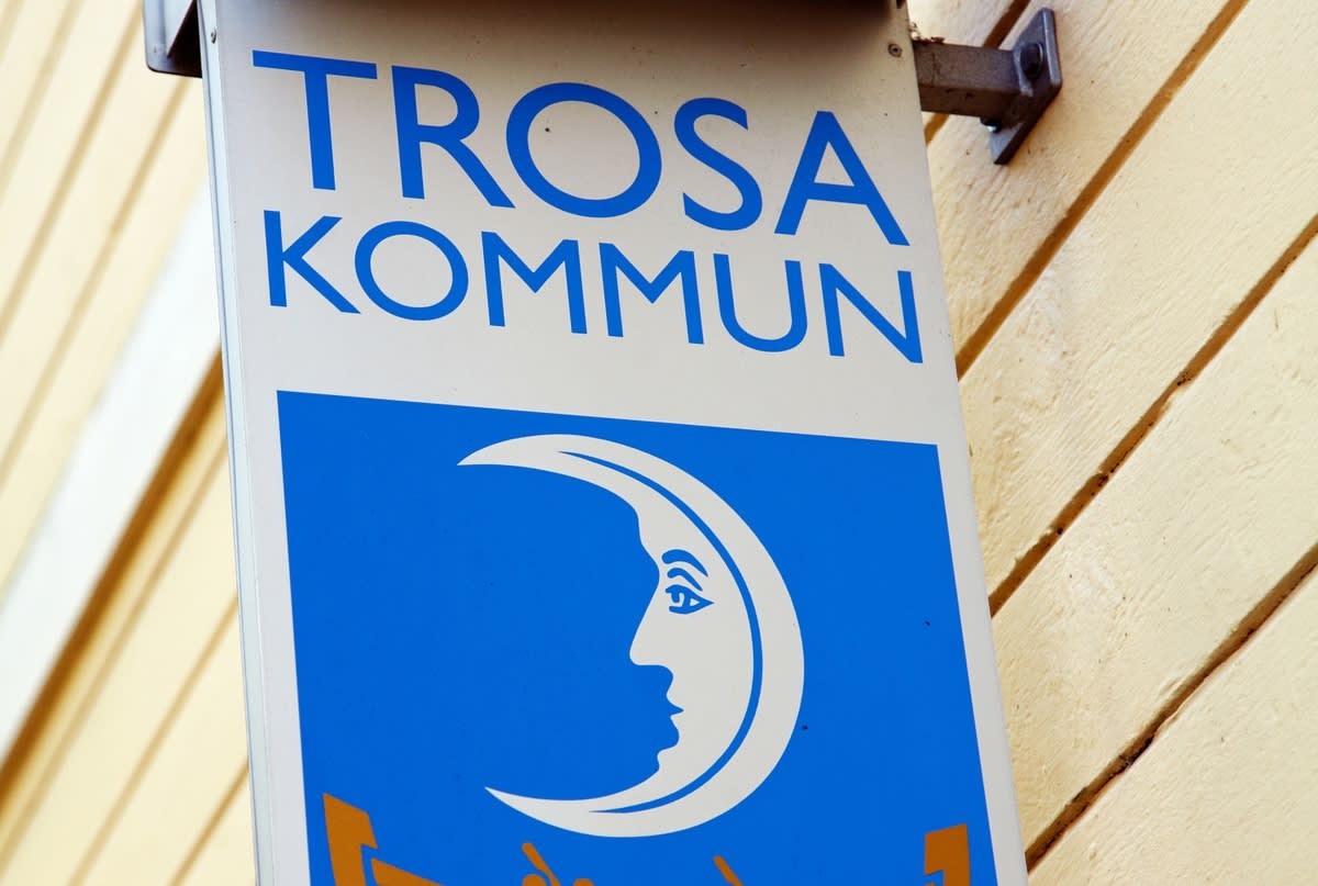 En nära bild av en vit fin skylt med texten "TROSA KOMMUN" i blå versaler överst. Under texten finns en blå ruta med en vit logotyp som föreställer en stiliserad måne med ett ansikte i profil. Skylten är monterad på en ljust gul träfasad med liggande