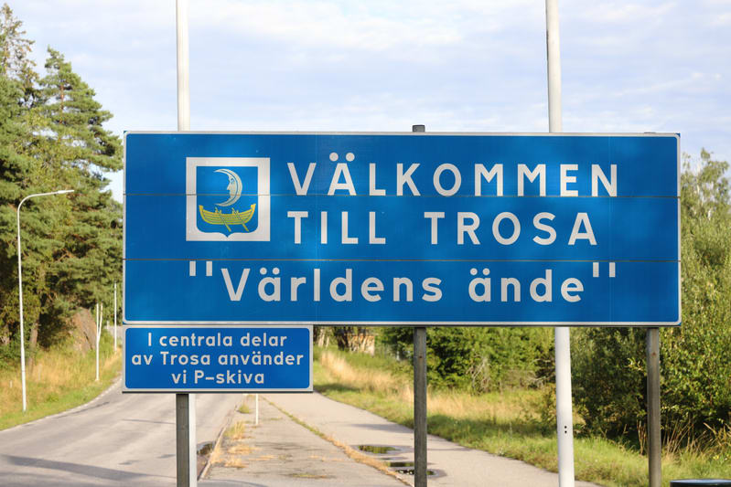 Bilden visar en blå välkomstskylt för Trosa, där texten "VÄLKOMMEN TILL TROSA "Världens ände"" är synlig i vitt. Ovanför texten finns Trosas kommunvapen med en halvmåne och en båt. Under den större skylten sitter en mindre blå skylt med texten "I cen