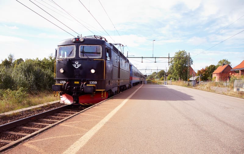 Bilden visar ett mörkt lokomotiv ståendes vid en järnvägsstation. Lokets front, med nummer 1359 och SJ:s logotyp, är vänd mot betraktaren och är svart med en röd och gul undersida. Lokomotivet är kopplat till flera passagerarvagnar som sträcker sig b