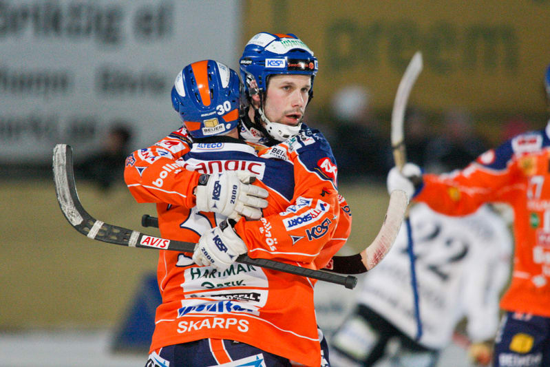 Bilden visar två bandyspelare i orangea och blåa dräkter som kramar varandra på isen. Båda bär hjälmar och den ena har nummer 30 på ryggen. I bakgrunden syns skymten av en tredje spelare och delar av en sarg med reklamtexter.