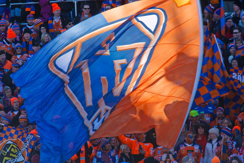 En stor blå och orange supporterflagga med ett mönster av bokstäver och en sköldliknande form täcker en del av bilden, framför en folkmassa av ansikten. Flaggans ena sida har en vit kant. Bakom flaggan syns ansikten på människor. Flera personer har o
