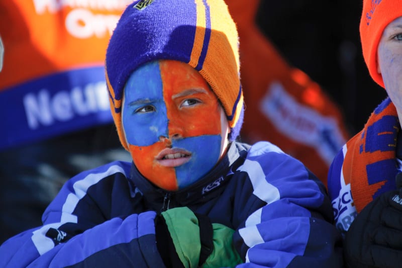 En närbild av ett barn vars ansikte är målat i blått och orange. Barnet bär en blå och orange mössa samt en blå jacka med vita detaljer. I bakgrunden syns suddiga skyltar med blå och orange färg. Ett annat barn syns delvis till höger.
