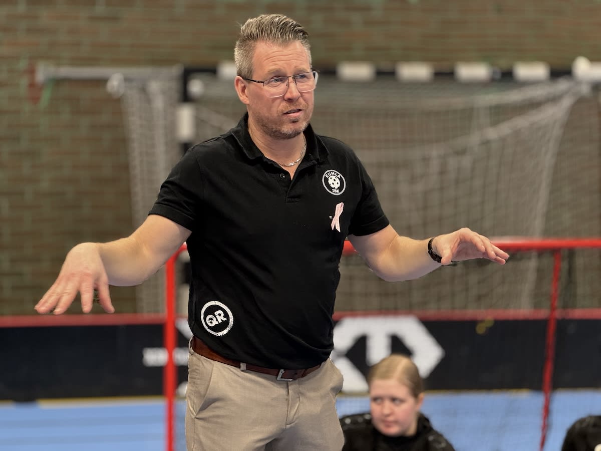 En man med glasögon, svart pikétröja och beigefärgade byxor står med utsträckta händer inomhus. Bakom honom syns handbolls-/innebandymål, en röd sarg, en tegelvägg och ett blått golv. En del av en annan person syns i förgrunden.