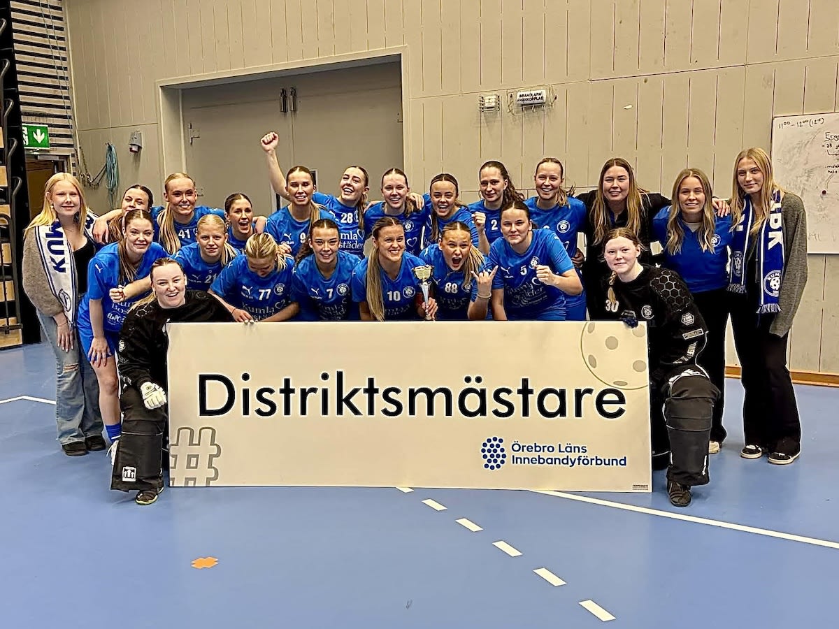 En grupp bestående av 21 personer deltar i en gruppbild i en idrottshall, där de håller upp en skylt med texten "Distriktsmästare" och logotypen för "Örebro Läns Innebandyförbund". De flesta personerna bär blå tröjor och några håller upp pokaler.