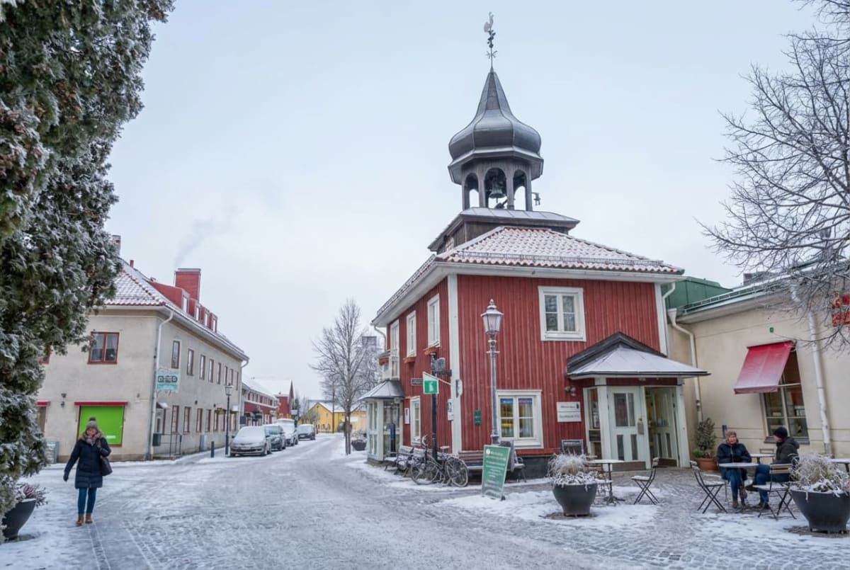En vinterbild av en snötäckt gata med flera byggnader och några personer. I förgrunden till vänster syns en kvinna gå, och till höger sitter två personer vid ett bord utanför en röd byggnad med ett klocktorn. Bilar står parkerade längs gatan i bakgru