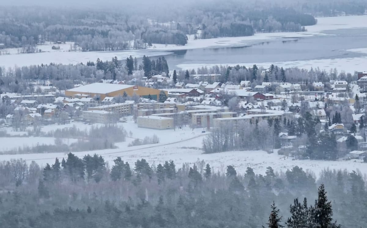 En översiktsbild av en vintertäckt bebyggelse vid ett vattendrag. Snö och frost täcker träd, mark och hustak. I bakgrunden syns ett delvis fruset vattendrag, omgivet av snötäckta skogar och en mindre by med bostadshus och större byggnader.