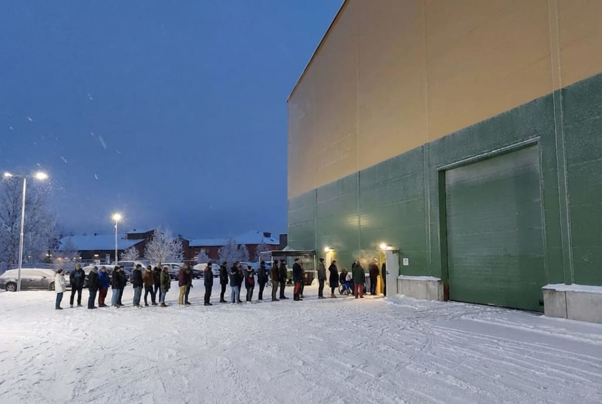 En grupp människor står i en kö utomhus, framför en stor byggnad, i ett vinterlandskap med snö på marken och träd. Det är mörkt med en blåaktig himmel och gatlyktor lyser upp området. En person i rullstol syns.