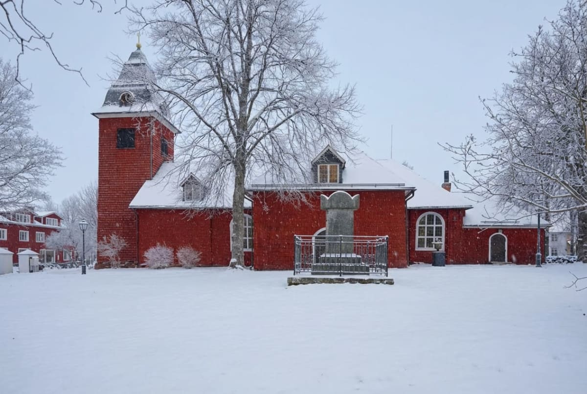 En röd byggnad med kyrktorn och snötäckta tak. Framför byggnaden finns ett snötäckt landskap med träd och en stenstaty omgiven av ett staket. I bakgrunden syns fler snötäckta byggnader och träd. Snön faller.