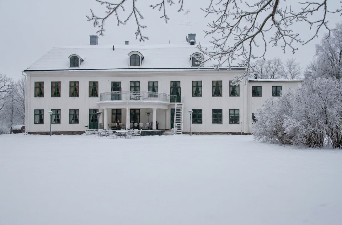 En stor, vit byggnad med flera fönster och ett snötäckt tak, framför byggnaden ligger ett snötäckt fält med möbler och utomhusbelysning. Delar av träd och buskar täckta med snö syns i bildens kanter och i bakgrunden.