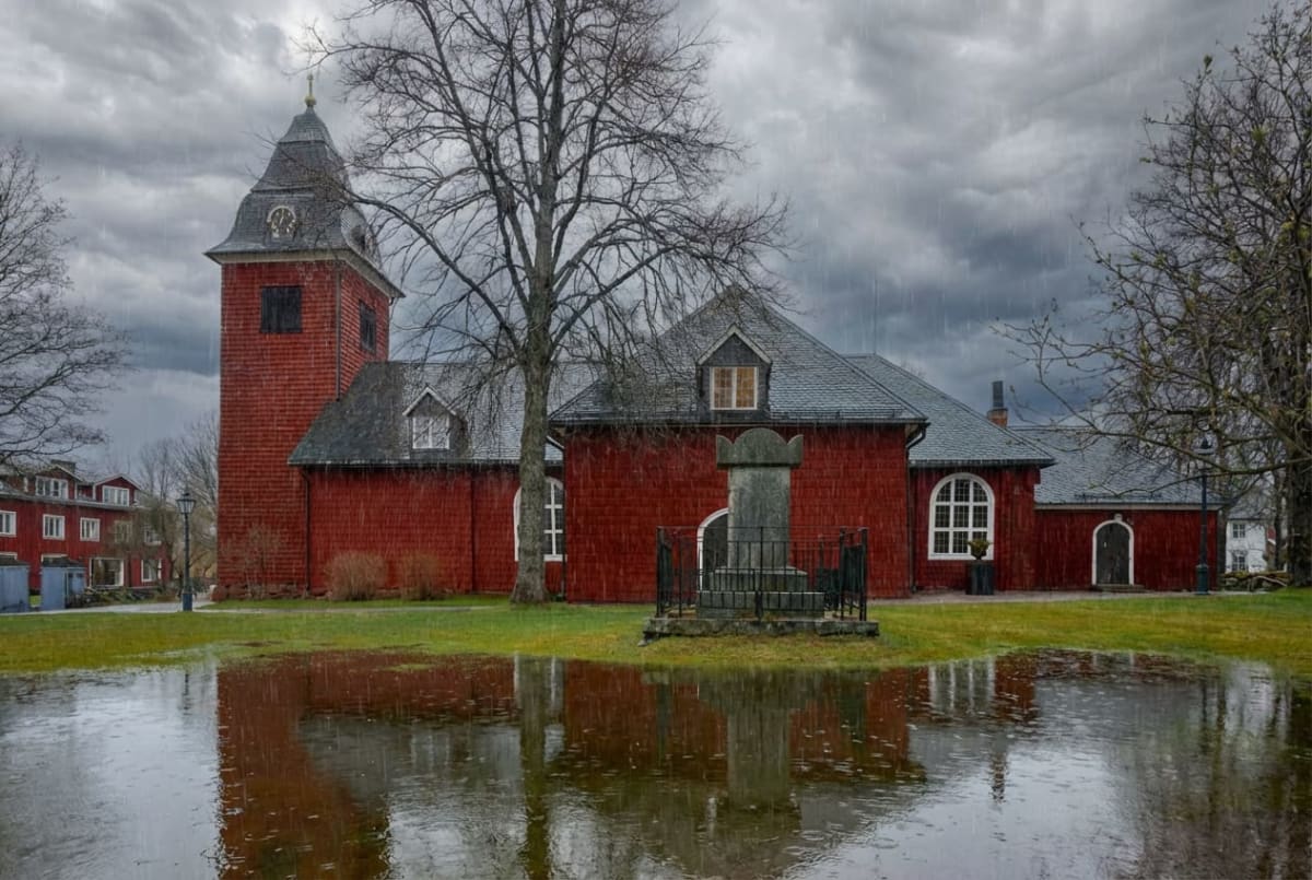 En röd träkyrka med ett grått tak syns i bilden, med en kyrktorn till vänster. Framför kyrkan finns en gräsmatta med stora vattenpölar som reflekterar kyrkan och himlen. Träd utan löv står runt kyrkan under en mulen och regnig himmel.