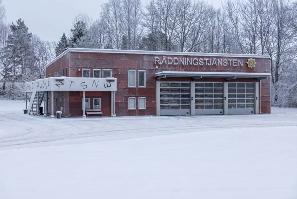 En röd byggnad, märkt med texten "RÄDDNINGSTJÄNSTEN", står i ett snötäckt landskap med träd i bakgrunden. Byggnaden har flera fönster och tre stora portar. Marken framför byggnaden är täckt av snö.