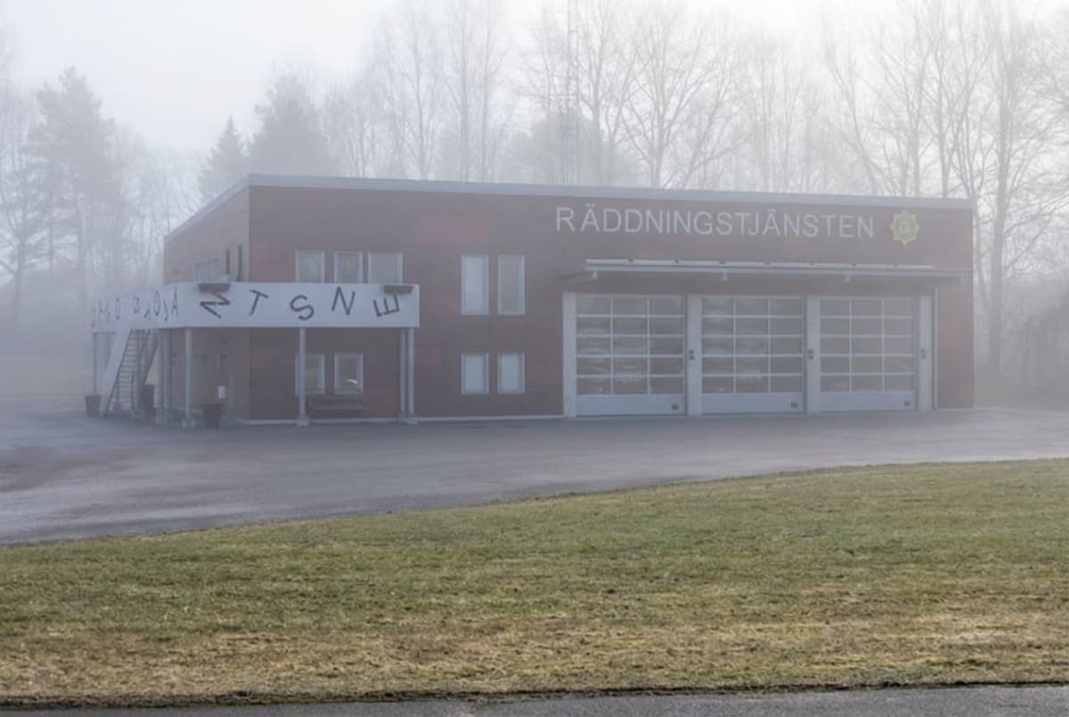 En röd byggnad med texten "RÄDDNINGSTJÄNSTEN" syns i den övre högra delen av bilden. På byggnaden finns även symboler och fönster, samt tre stora vita portar. Framför byggnaden finns en grön gräsmatta, en asfalterad yta och en trappa till vänster med