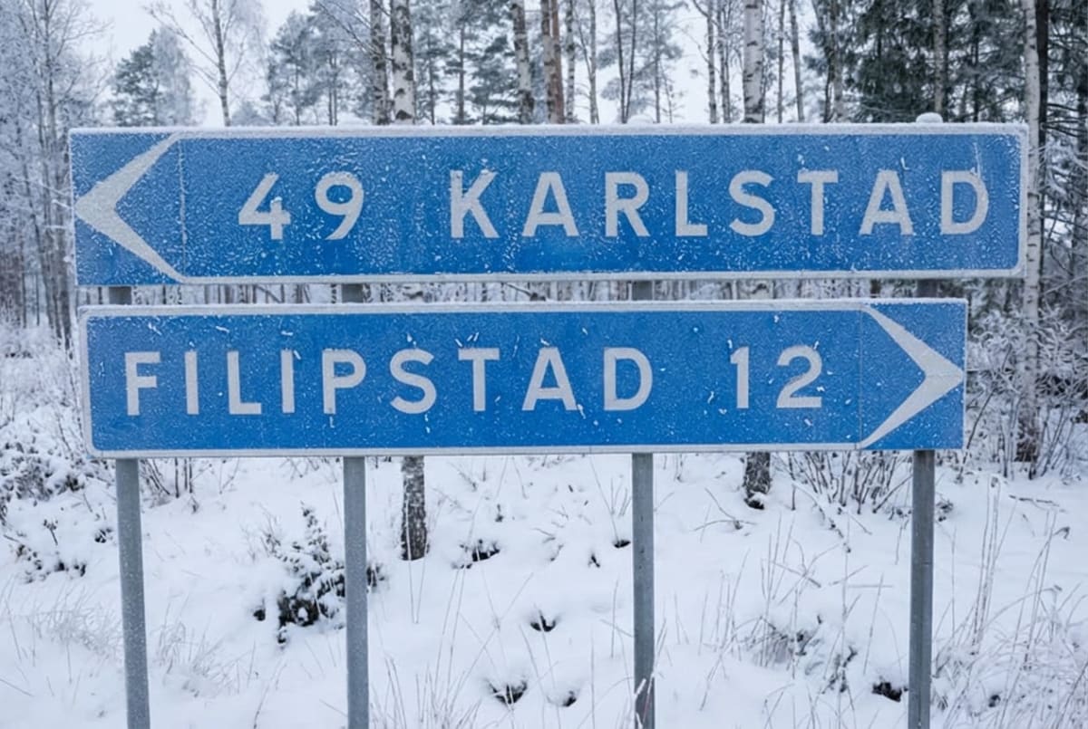En blå skylt med vita pilar och texten "49 KARLSTAD" och "FILIPSTAD 12". Skylten är täckt av rimfrost och står i ett snötäckt landskap med barr- och lövträd i bakgrunden.