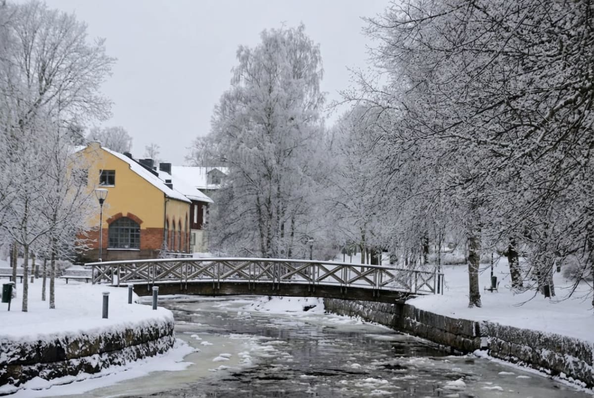 En vinterbild som visar en bro över ett delvis istäckt vattendrag. På vardera sida av vattendraget finns snötäckta träd och marken är täckt av snö. I bakgrunden syns gul- och rödaktiga byggnader. Himmelen är grå och mulen.