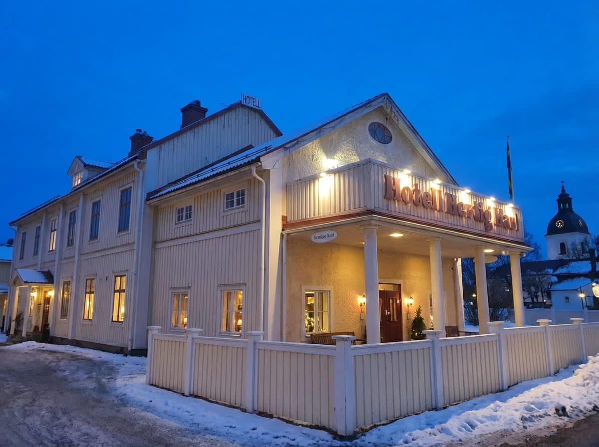 En byggnad med fasad i ljusa träpaneler och puts, som ser ut att vara ett hotell. Byggnaden har flera fönster med lysande ljus inifrån och en upplyst entré. En snötäckt mark och en vit staket syns i förgrunden, och ett kyrktorn skymtar i bakgrunden m