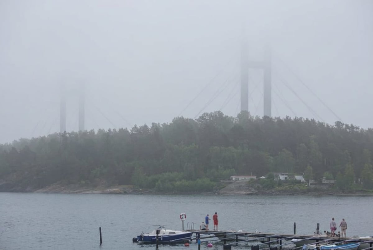 En bild tagen utomhus under dimmiga förhållanden som visar en bro, en skogklädd ö och en brygga med människor och båtar i förgrunden. Brokonstruktionen är delvis skymd av dimma.