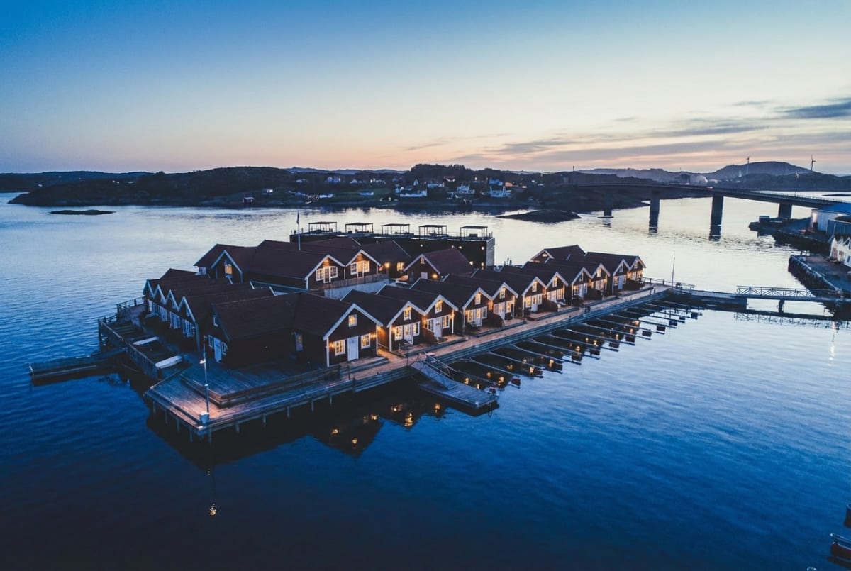 En översiktsbild av en rad sammanbyggda sjöbodar eller småhus, placerade på en brygga eller flytande plattform i vatten. Ljussken från fönstren reflekteras i vattnet. I bakgrunden syns landskap, en bro, byggnader och en himmel i skymningen.