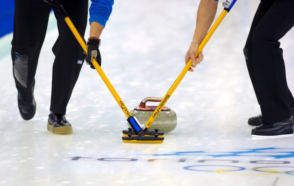 Två personer ses från midjan neråt hållandes i var sin gul curlingborste som de sätter ner mot isen, nära en curlingssten, även den på isen. På isen syns även flertalet olympiska ringar i olika färger.