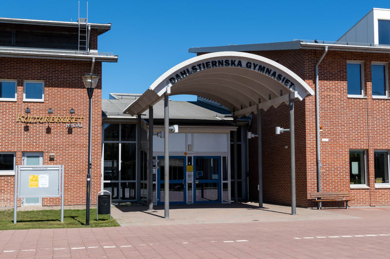 En fasadbild av en större byggnad med tegelväggar och ett täckt ingångsparti. Ovanför ingången står texten "DAHLSTIERNSKA GYMNASIET". Till vänster finns en skylt med texten "KulturBruket På Dal" och en anslagstavla. Till höger om ingången finns en bä