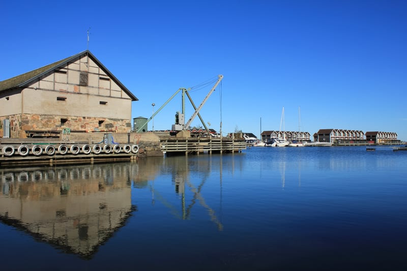 En bild visar en marina med en stor, ljus byggnad till vänster och en kaj med upphängda bildäck. En lyftkran syns i mitten av bilden, och till höger ligger flera segelbåtar förtöjda framför en rad byggnader med röda tak. Vattnet reflekterar båtarna,