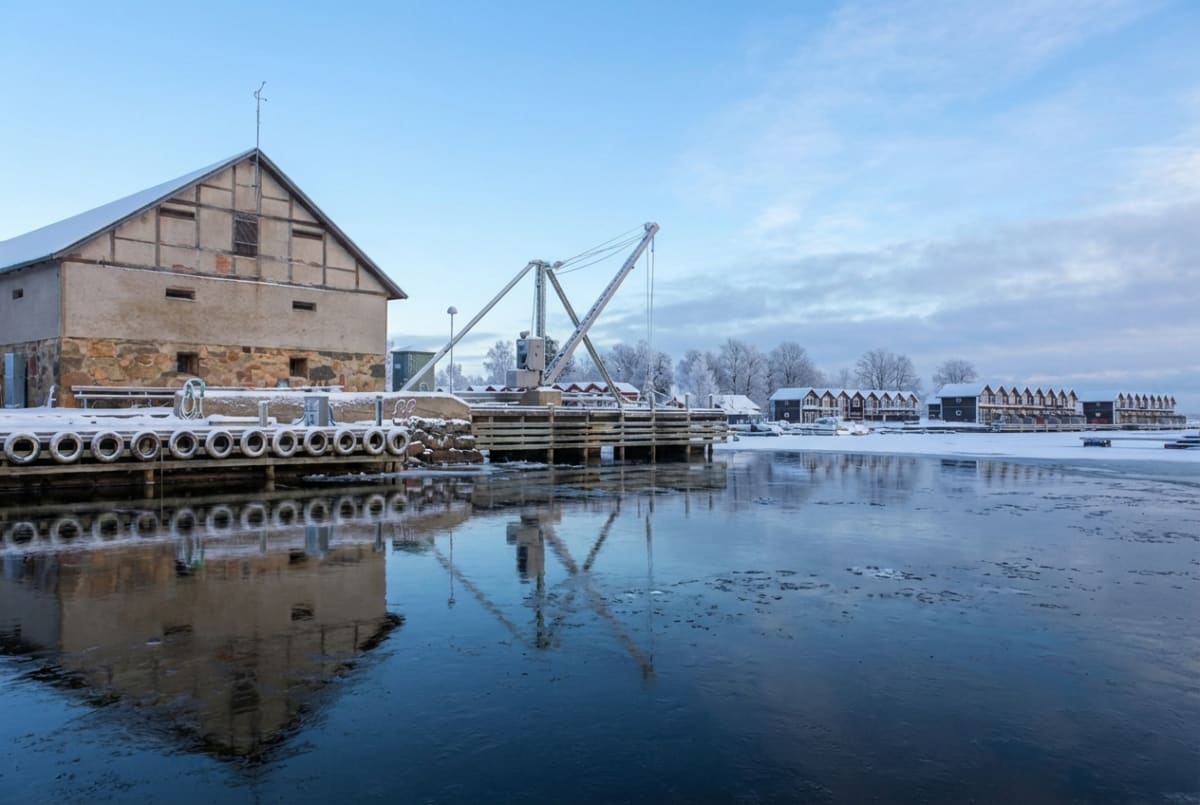 En vinterbild från en kustmiljö med en byggnad, en kran och en brygga. Vattnet är delvis islagt och har reflektioner. I bakgrunden syns snöklädda träd och byggnader längs vattenkanten under en ljusblå himmel med moln.