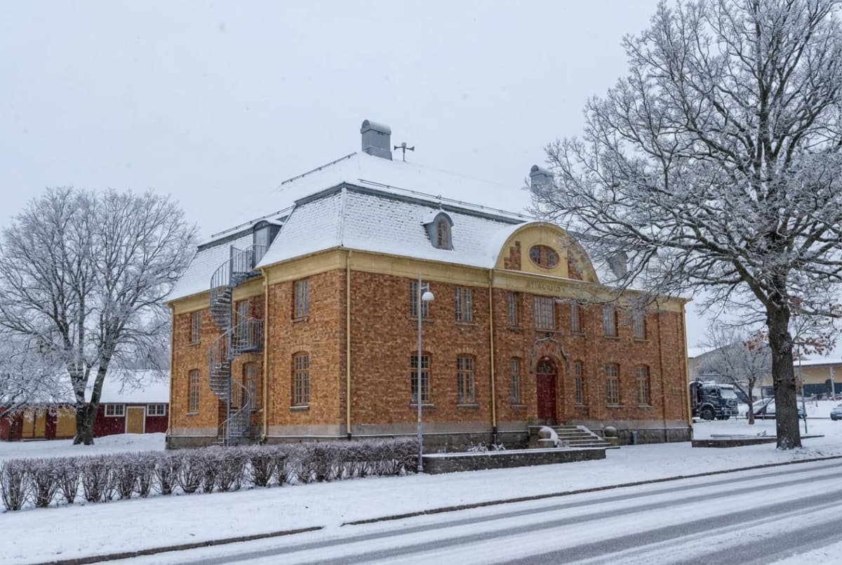 En stor tegelbyggnad med sadeltak täckt av snö, en röd dörr och flera fönster. Byggnaden har en gul avsats under taket och en spiraltrappa i metall på ena sidan. Träd utan löv, täckta av snö, syns i förgrunden och bakgrunden. En snötäckt väg syns i f