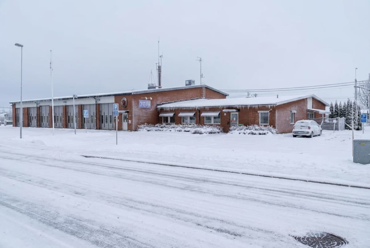 En röd tegelbyggnad med flera garageportar och ett långsträckt, täckt tak ses under en snöig vinterdag. Ett vitt fordon är parkerat bredvid byggnaden. Marken och vägen framför är täckta av snö.