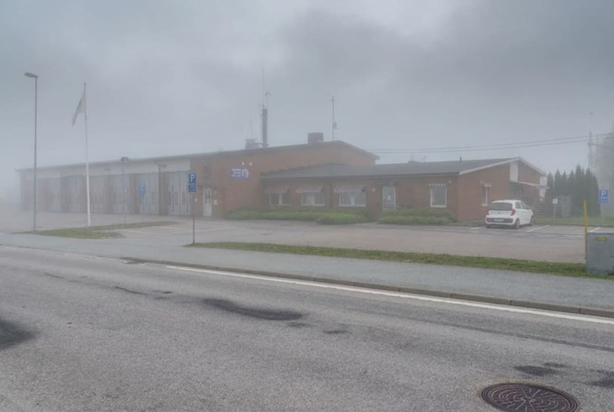 En bild tagen utomhus under en dimmig dag som visar en tegelbyggnad med flera garageportar. En vit personbil står parkerad utanför garaget. En svensk flaggstång med flagga syns till vänster om byggnaden, och närmast betraktaren finns en väg med en br