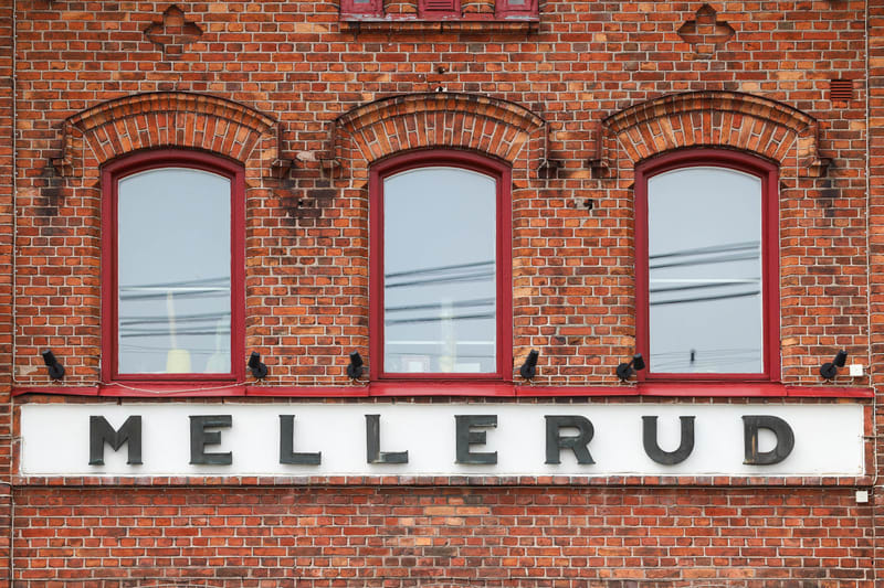 En fasad av rött tegel med tre rödramade fönster. Under fönstren sitter en vit skylt med texten ”MELLERUD” i svarta versala bokstäver. På tegelväggen syns även flera svarta fängar för externa armaturer.