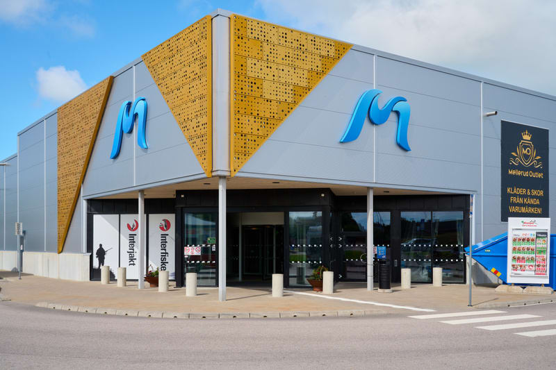 En modern byggnad med en butiksfasad dominerar bilden. Byggnaden har gråa metallpaneler och en dekorativ gulbrun perforerad fasaddetalj. Ett stort blått "M" är monterat på fasaden. Ingången består av glasdörrar, och framför den finns ett asfalterat o