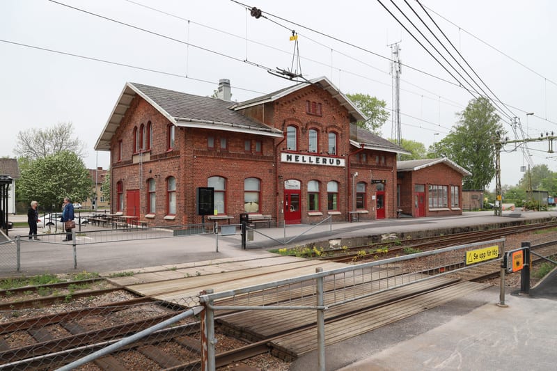 En röd tegelbyggnad med texten "MELLERUD" på framsidan, framför vilken det finns järnvägsspår och en perrong. Två personer står på perrongen till vänster i bilden. I bakgrunden syns träd, bostadshus och en mobilmast. En gul skylt med texten "Se upp f