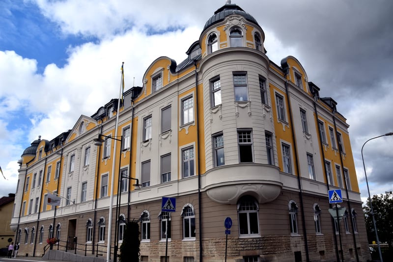 En stor, äldre byggnad med gul och ljus fasad i flera våningar under en delvis molnig himmel. Byggnaden har många fönster och en rundad hörnsektion med ett kupolformat tak. Två gatuskyltar syns framför byggnaden, varav en är en övergångsstället-skylt