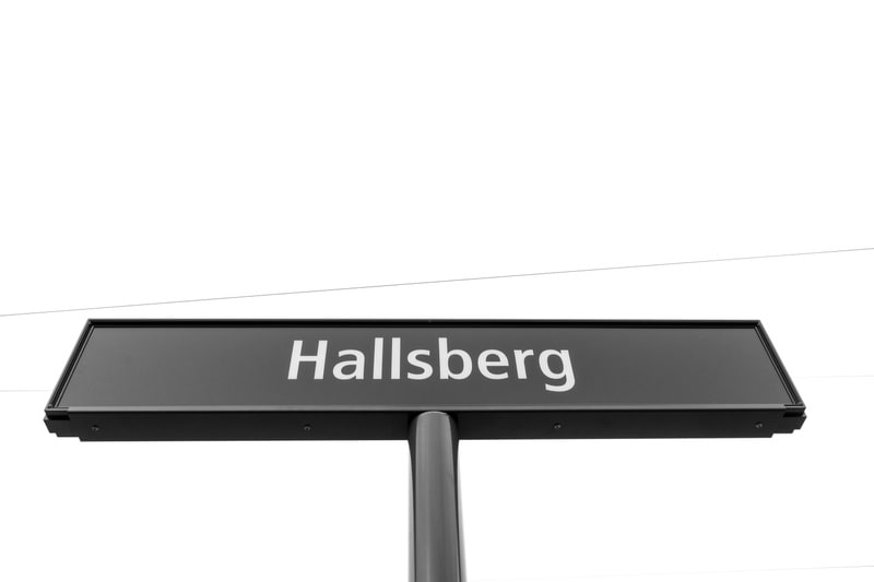 En svart skylt med texten "Hallsberg" i ljusa, versala bokstäver. Skylten är monterad på en stolpe och synes mot en ljus, nästan vit bakgrund. Tunna linjer syns i det övre vita fältet.