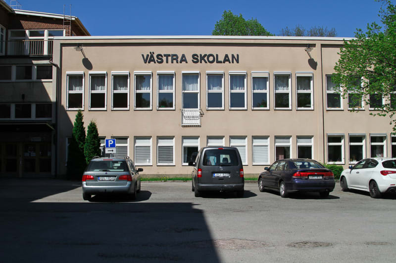 En beige byggnad med texten "VÄSTRA SKOLAN" på fasaden. Framför byggnaden finns en asfalterad parkeringsplats med flera bilar. Träd med gröna löv syns i bildens övre del.
